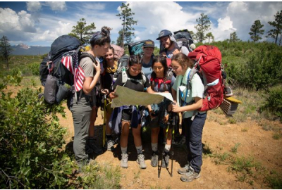 A group of seven female Scouts read a map on a dirt trail on a summer day. 