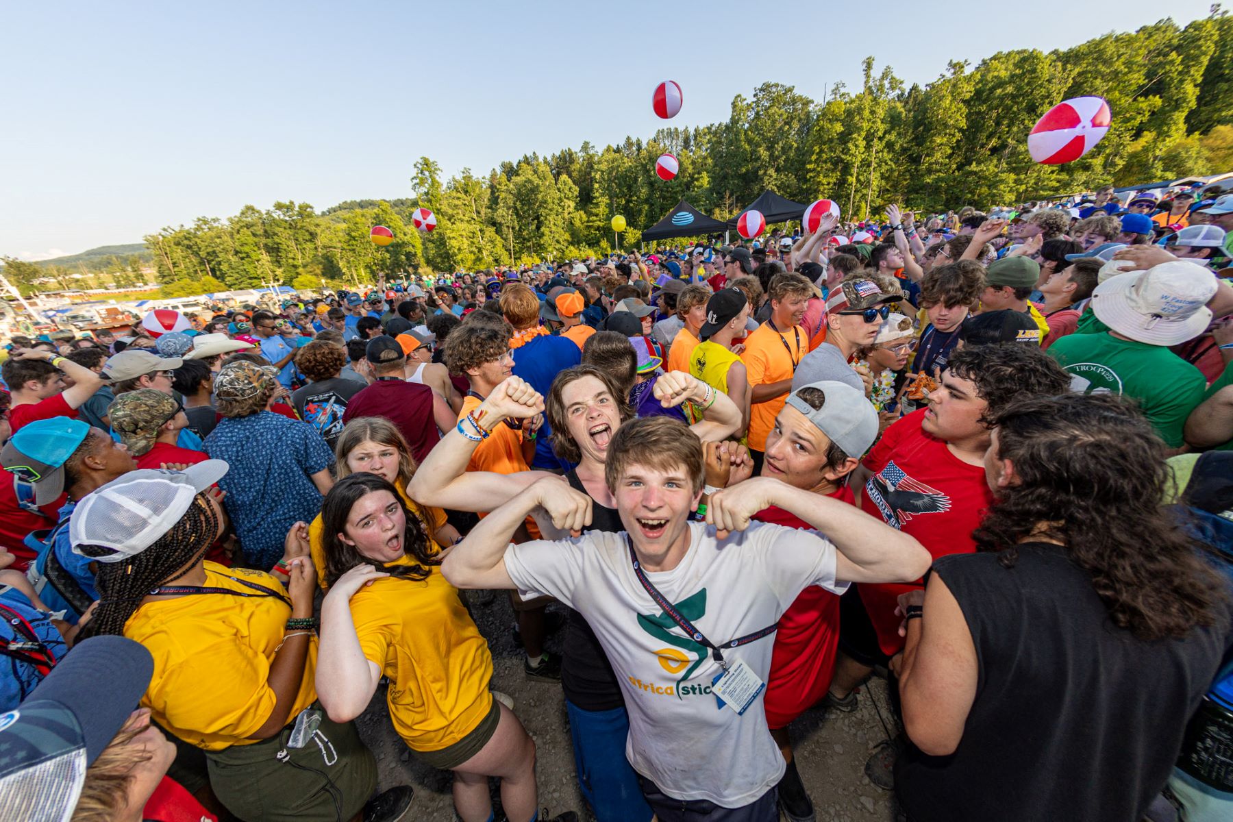 Crowd of Scouts celebrating at the National Jamboree, smiling and cheering with beach balls in the air and tents and trees in the background.