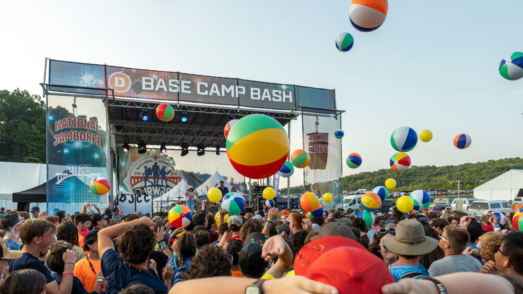 Scouts gather in front of the Base Camp Bash stage at the National Jamboree, surrounded by colorful beach balls and outdoor tents with trees in the background.