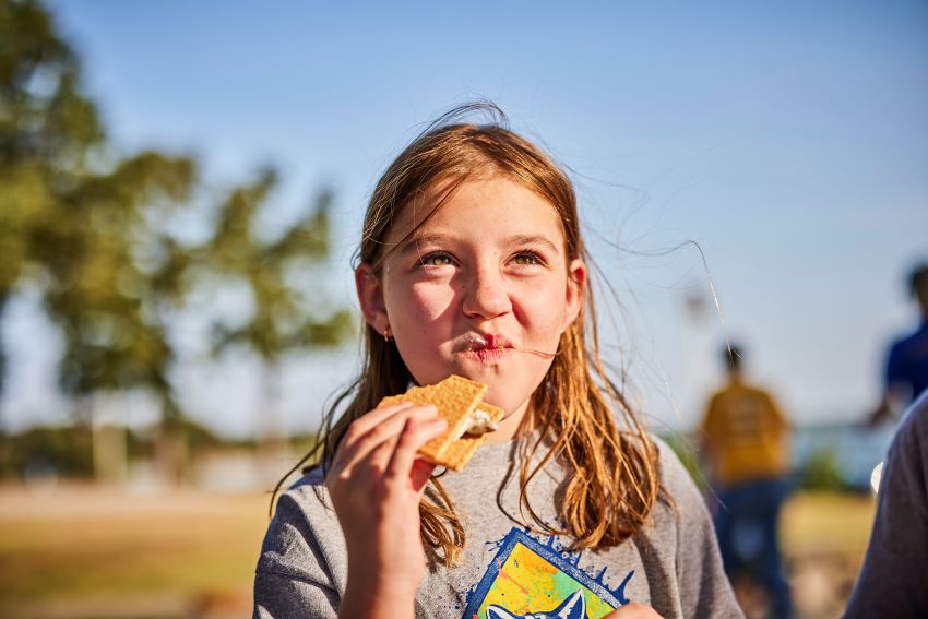 A young girl wearing a gray Cub Scout t-shirt enjoys a s'more outdoors, with a happy expression on her face and a background of trees and fellow Scouts.