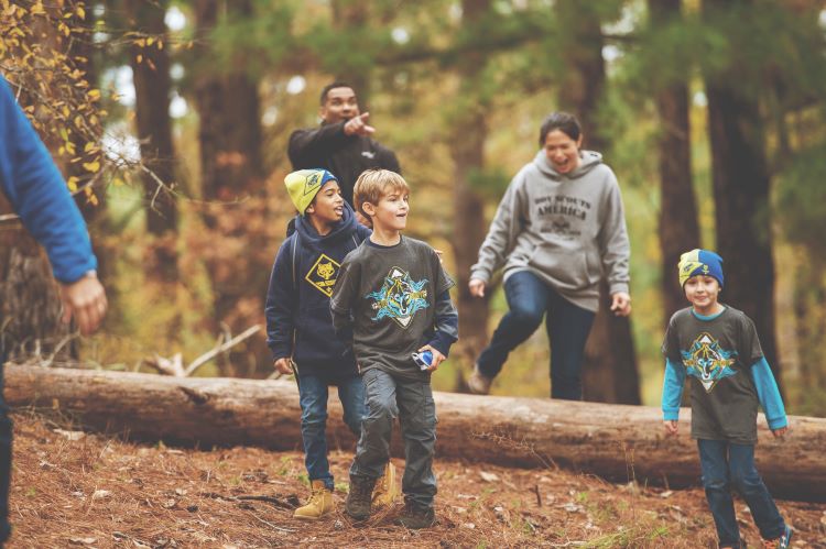 Three young children and two adults walking over a fallen tree in the woods during fall.