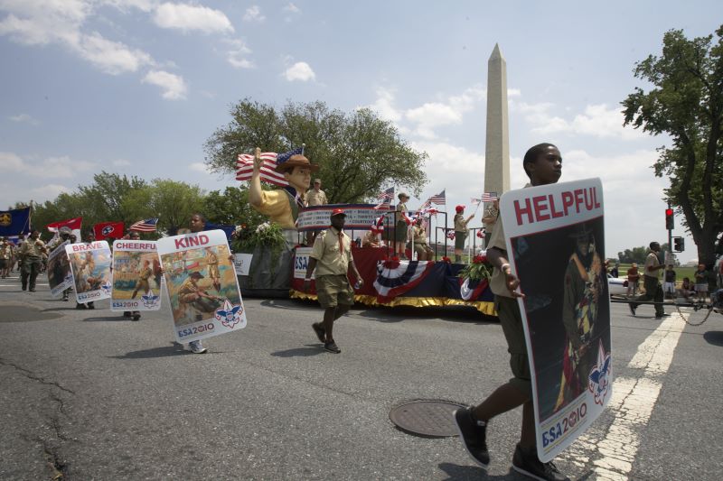 Scouts march in a parade near the Washington Monument, proudly carrying signs representing the Scout Law during a national celebration.