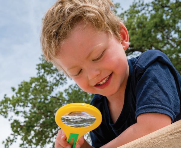 A young boy in a Cub Scout uniform smiles as he looks through a magnifying glass, exploring his surroundings on a sunny day with trees in the background.