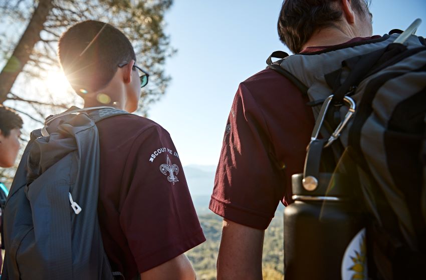 Two Scouts wearing matching maroon shirts hike through a scenic outdoor area with backpacks and reusable water bottles, ready to participate in a conservation or environmental stewardship project.