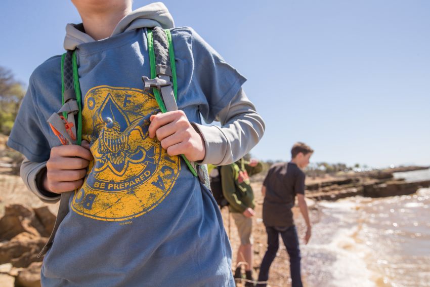 A Scout wearing a blue Scouting t-shirt and backpack stands near the water, ready for an outdoor cleanup mission. Other Scouts walk along the rocky shoreline in the background, participating in an environmental conservation activity.